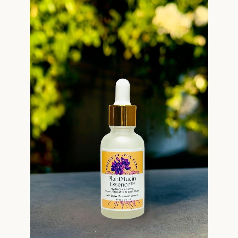 May include: A clear glass bottle with a white dropper and gold cap, labeled "Plant Mucin Essence." The label is yellow with purple floral graphics and text that reads "Hydrates + Firms" and "1 fl oz / 30 ml."