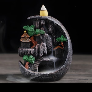 May include: A black ceramic incense burner shaped like a crescent moon with a mountain scene. The burner features a small house, trees, and a waterfall. The incense cone is placed on top of the mountain.