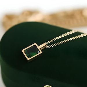 May include: A gold necklace featuring a rectangular, emerald-green gemstone pendant. The pendant is set in a gold frame and suspended from a delicate chain. The necklace is displayed on a dark green velvet surface.