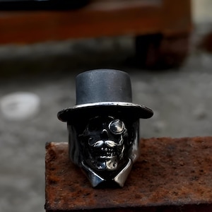 May include: A silver ring featuring a skull design wearing a top hat, monocle, and a mustache. The ring has a detailed, gothic aesthetic. The ring is displayed on a rusty metal surface.