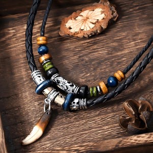 Puede incluir: Un collar artesanal con un cordón de cuero negro trenzado. El collar presenta cuentas de madera, detalles metálicos plateados y un colgante con forma de diente. El collar se exhibe sobre una superficie de madera.