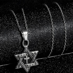 May include: Silver-toned necklace featuring a Star of David pendant with a cross at its center. The pendant has an intricate design and hangs from a delicate chain. The necklace is displayed on a textured black surface.