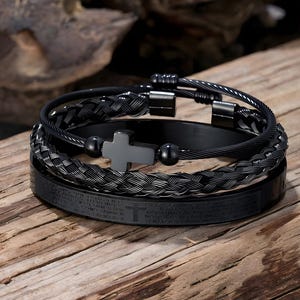 Può includere: Un set di braccialetti neri con un ciondolo a forma di croce. I braccialetti hanno diversi design, tra cui stili intrecciati e a cavo. I braccialetti sono presentati su una superficie di legno.