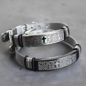 Puede incluir: Dos pulseras plateadas con detalles negros y una banda de malla. Cada pulsera presenta una cruz y texto grabado. El texto incluye la frase "Padre nuestro que estás en el cielo".
