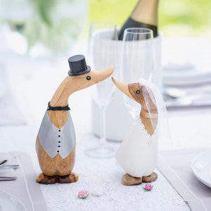 May include: Wooden duck figurines, one in a tuxedo with a top hat and the other in a wedding dress and veil. The ducks are positioned as if kissing, with a champagne bottle and glasses in the background. Decorative wedding theme.