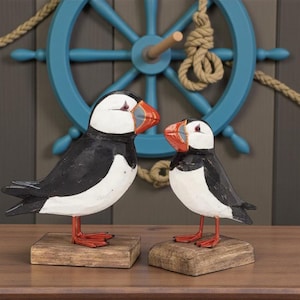 Puede incluir: Dos figuras de frailecillos de madera. Las aves tienen cuerpos blancos y negros, patas rojas y picos naranjas. Están montadas sobre pequeñas bases cuadradas de madera. Un timón azul y una cuerda decoran el fondo.