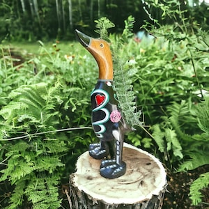 May include: A decorative wooden duck figurine with a brown neck and head, a black body adorned with a red heart and floral designs, and blue feet. The duck is standing on a tree stump, surrounded by green foliage.