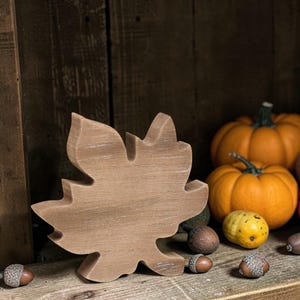 May include: A wooden leaf decoration in a light brown color, with visible wood grain, sits on a wooden shelf. The background features orange pumpkins, acorns, and a dark wooden backdrop, creating a fall-themed display.