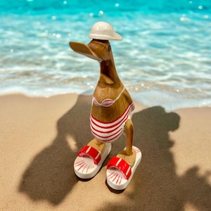 Puede incluir: Una figurita de pato de madera con un sombrero blanco y un traje de baño a rayas rojas y blancas. El pato está de pie en una playa de arena con agua azul de fondo.
