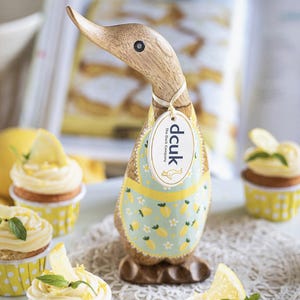 May include: Wooden duck figurine with a light brown body and a wooden head. The duck wears a light blue apron with yellow lemons and white flowers. A tag reads "dcuk The Duck Company". Cupcakes with lemon slices and mint leaves are in the foreground.