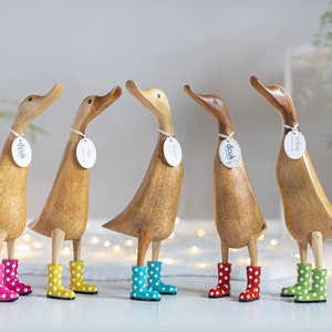 Könnte beinhalten: Fünf hölzerne Entenfiguren, die bunte, gepunktete Gummistiefel tragen. Die Enten stehen auf einer weißen Oberfläche mit einer Lichterkette im Hintergrund.