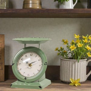 May include: A vintage-style, mint green scale clock with a distressed finish. The clock face has black numerals and hands, with the text "OLD TOWN GOODS" at the bottom. A small vase with yellow flowers sits to the right, adding a touch of color to the scene.