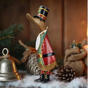 May include: A wooden duck figurine dressed in a red and green military-style uniform and a black top hat. The duck is wearing black boots and has a tag hanging from its neck. The figurine is a decorative item, possibly for the holidays.