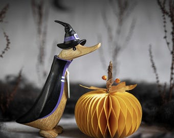 Puede incluir: Una figurita de pato de madera vestida de bruja con un sombrero morado y negro y una capa negra. El pato está de pie junto a una calabaza amarilla en forma de panal.