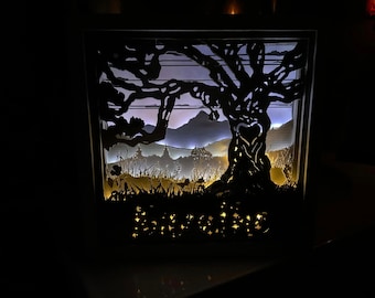 Könnte beinhalten: Ein Papier-Schnittbild, das einen Baum mit einer herzförmigen Öffnung im Stamm zeigt. Der Baum wird von hinten beleuchtet und wirft einen warmen Schein auf die umliegende Landschaft. Die Szene umfasst Berge, Gras und einen kleinen Vogel.