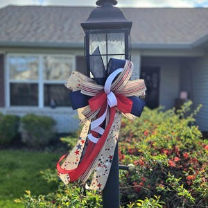 May include: A black lamp post with a red, white, and blue bow tied around it. The bow is made of ribbon with stars and stripes. The lamp post is in front of a house.