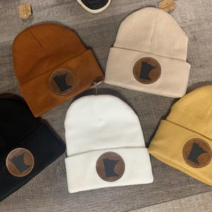 Puede incluir: Cinco gorros de punto de diferentes colores con un parche de cuero marrón que presenta el contorno del estado de Minnesota. Los gorros están dispuestos sobre una superficie de madera.
