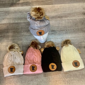 May include: Five knit winter hats with faux fur pom poms. Each hat features a brown leather patch with the outline of the state of Minnesota. The hats are in the colors gray, beige, pink, black, and white.
