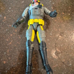 May include: A G.I. Joe action figure featuring a gray tactical vest, yellow utility belt, and yellow leg accents. The figure wears a black helmet with a red visor, black boots, and black gloves, with a red Cobra logo on the chest.