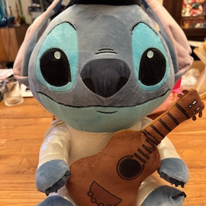 May include: A plush Stitch doll wearing a black hat and a sparkly white and blue outfit, holding a brown ukulele. The doll has large black eyes, blue accents, and is sitting on a wooden surface.
