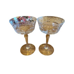 Puede incluir: Dos copas de champán con diseños florales grabados. Las copas tienen una base redonda, un tallo retorcido y un cuenco ancho. El cristal es transparente con un ligero tinte ámbar.