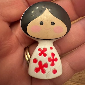 May include: A vintage doll-shaped figurine with a black head, rosy cheeks, and a white dress adorned with red flower designs. The figurine is held in a hand, showcasing its small size and detailed features. A charming collectible.