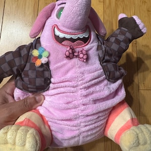 Può includere: Un peluche con la pelle rosa, una grande proboscide e un ampio sorriso che mostra i denti bianchi. Indossa una giacca a scacchi marrone e nera, un piccolo papillon a pois e una spilla a fiore multicolore. Le gambe presentano strisce.
