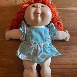 Könnte beinhalten: Eine Puppe mit weichem Stoffkörper und geformtem Kunststoffkopf, die leuchtend orangefarbenes Wollhaar mit einem gemusterten Haargummi trägt. Die Puppe ist mit einem hellblauen Kleid mit Rüschenärmeln und einer weißen Blumenborte bekleidet.