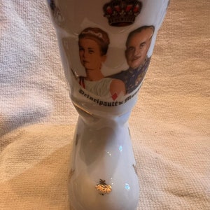 Może przedstawiać: Biała ceramiczna wazon w kształcie buta z pozłacanym wykończeniem. Wazon przedstawia portret pary z koroną nad nimi. Na wazon widnieje napis "Principauté de Mon".