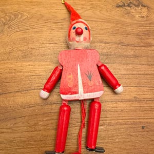 May include: A vintage wooden pull-string toy in the shape of a gnome. The gnome has a red pointed hat, a white beard, and a red outfit with painted details. The arms and legs are jointed, and the toy is suspended by a gold string.