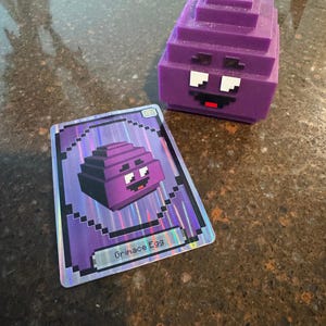 Puede incluir: Una figura púrpura pixelada de Grimace Egg y una tarjeta coleccionable a juego. La figura tiene un diseño en bloques con ojos blancos y boca roja. La tarjeta presenta el mismo diseño con el texto "Grimace Egg".