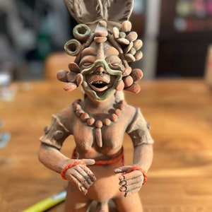 May include: A terracotta figurine of a kneeling figure with a green and brown face mask, wearing a beaded necklace and a red rope around the waist. The figure has a headdress with flowers and a large, round, green and brown object on top.