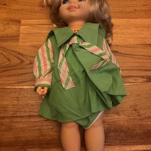 May include: A vintage doll with blonde hair and blue eyes, dressed in a green dress with a matching bow and a striped shirt. The doll is lying on a wooden surface.