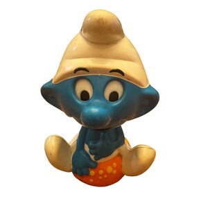 Puede incluir: Una figura vintage de los Pitufos, con cara y cuerpo azules, gorro blanco y postura sentada. La figura sostiene una bola naranja con puntos amarillos. El juguete tiene un atractivo clásico y nostálgico.