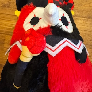 Puede incluir: Una muñeca de bufón de peluche con una combinación de colores negro y rojo. La muñeca tiene una cara blanca con ojos negros y un cuerno blanco. El sombrero del bufón es a cuadros rojos y negros. La muñeca tiene un cuerpo rojo y negro.