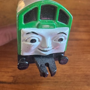 May include: A green and white toy train with a smiling face. The train has a green roof and windows, a white face with black eyes, and a black coupling. The toy is held by a hand.