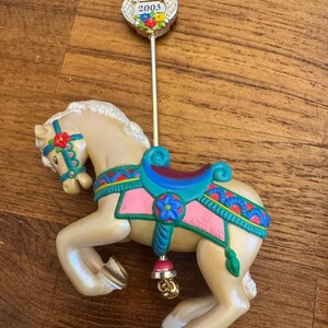 Puede incluir: Adorno de caballo de carrusel crema, adornado con una silla de montar turquesa, detalles en rosa y detalles florales. El adorno tiene una campana dorada y una parte superior decorativa con el año 2003.