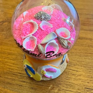 May include: A clear glass snow globe with a pink sand base, seashells, and the words "Miami Beach" on the base. The base features a yellow and blue flip-flop and a pink hat. The globe is filled with pink sand and various seashells.