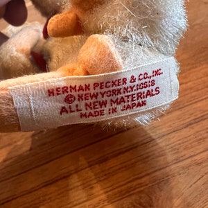 May include: A white plush toy with a red mouth and orange feet. The toy has a white label sewn on the bottom with the text "HERMAN PECKER & CO., INC. NEW YORK N.Y. 10018 ALL NEW MATERIALS MADE IN JAPAN."