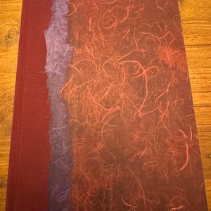 May include: A rectangular book with a dark red spine and a cover featuring a vertical strip of textured purple paper next to a larger section of dark red paper with an embedded pattern of thin, fibrous red strands throughout the surface.