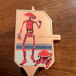 May include: A red and white enamel pin featuring two red robots. The larger robot is standing with its arms outstretched. The smaller robot is crouching. The text "PIT DROID" is printed on the pin.
