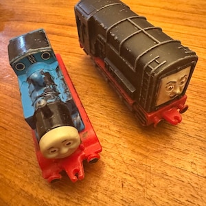 May include: Two toy train engines. One is black with red detailing and a face. The other is blue and red with a face. Both are on a wooden surface.