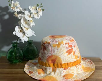 May include: A wide-brimmed sun hat with a white background and a sunflower print in shades of orange and yellow. The hat has a yellow band around the crown.