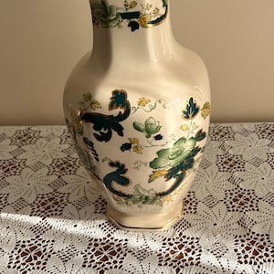 Puede incluir: Jarrón de cerámica color crema con la parte superior acampanada y base ancha, adornado con motivos florales verdes y amarillos. El borde tiene un borde verde oscuro con detalles dorados. El jarrón está sobre un mantel de encaje blanco.