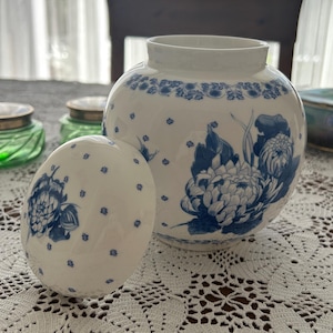 May include: A white ceramic jar and lid with blue floral designs and polka dots. The jar is round with a wide opening, and the lid is egg-shaped. The jar and lid are on a white lace tablecloth.