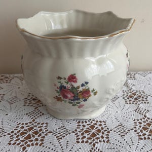 Puede incluir: Jardinera de cerámica color crema con borde festoneado y ribete dorado. La jardinera presenta un diseño floral con flores rosas, azules y amarillas. Se encuentra sobre un mantel de encaje blanco, de estética vintage.