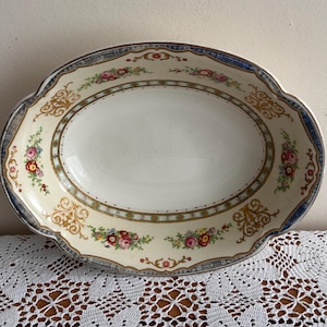 以下が含まれることがあります： クリーム色の内側と装飾的なボーダーが付いた楕円形のサービングディッシュ。ボーダーは、青とグレーのトリムが付いた花柄と金色のスクロールワークのデザインが特徴です。白いレースのテーブルクロスの上に置かれています。