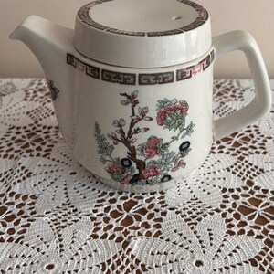 May include: A white ceramic teapot with a lid, decorated with a floral pattern in pink, brown, and green. The teapot has a spout and handle, and is placed on a white lace tablecloth. A traditional tea item.