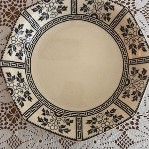 May include: An octagonal cream plate with a black floral and geometric border design. The plate has a central cream area and is set on a white lace tablecloth.