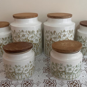 Puede incluir: Conjunto de seis botes de cocina de cerámica blanca con tapas de madera, decorados con un diseño floral verde y marrón. Dos botes están etiquetados como "TEA" y "COFFEE".